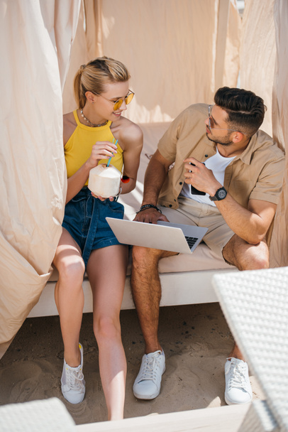 giovane coppia in occhiali da sole utilizzando computer portatile e sorridendo l'un l'altro mentre seduti insieme in bungalow sulla spiaggia
 - Foto, immagini