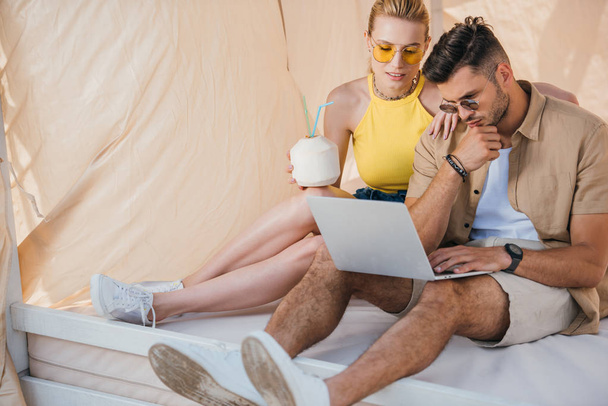 young couple in sunglasses using laptop while sitting in bungalow - Foto, imagen