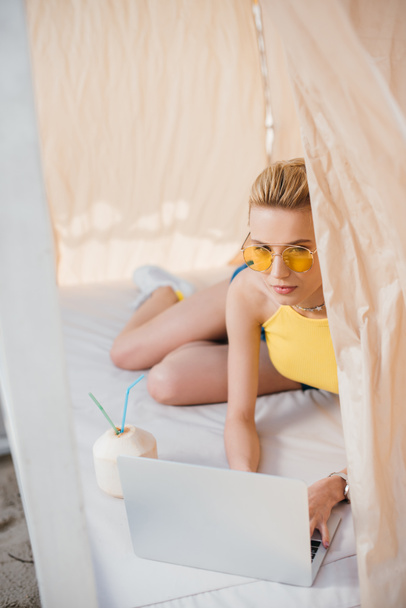 bela jovem mulher em óculos de sol usando laptop enquanto deitado em bangalô
 - Foto, Imagem