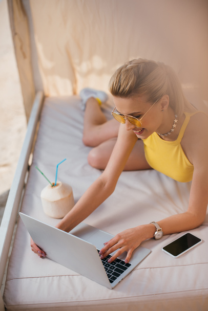 high angle view of smiling beautiful girl in sunglasses using laptop while resting in bungalow - Foto, Bild
