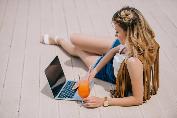 hermosa mujer joven usando el ordenador portátil y sosteniendo el vaso con cóctel
  - Foto, Imagen