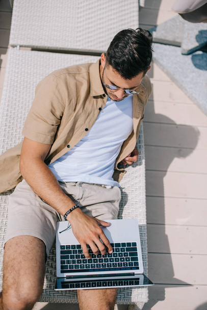high angle view of young man in sunglasses lying on chaise longue and using laptop - Φωτογραφία, εικόνα