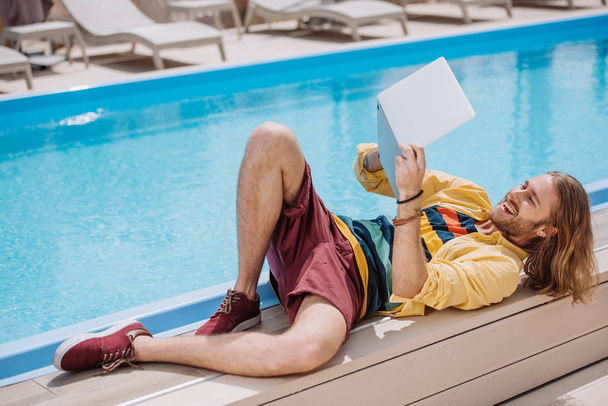 uśmiechnięty mężczyzna młody za pomocą laptopa leżąc w pobliżu basenu - Zdjęcie, obraz