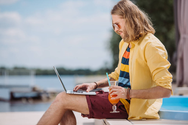 Widok z boku młodego człowieka w okulary wciskając szkła koktajl i laptop na plaży - Zdjęcie, obraz