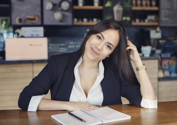 Beautiful hispanic young woman in suit working in the cafe writing in a copybook o diary, smiling looking at camera - Foto, Imagem