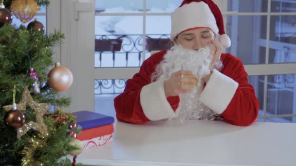Bored man in Santa Claus suit with fake beard sit on the table and drinking a glass of strong alcohol. Male is sad and tired. - Imágenes, Vídeo