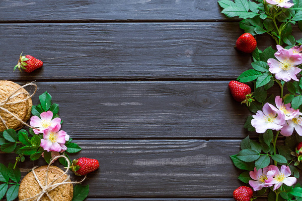 oatmeal cookies, strawberry and flowers with leaves on a black wooden background, background for text - Foto, imagen