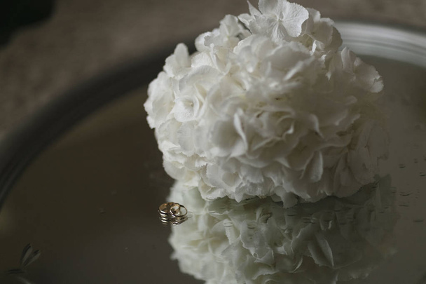 A bouquet of white hydrangeas and wedding rings with drops of water around lie on a glass table. Close-up horizontal photo - Foto, Bild