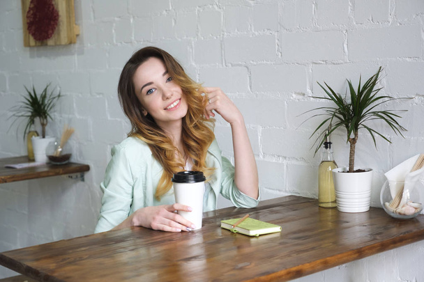Beautiful young girl sitting at the table with coffee and smiling looking at the camera, straightening hair. Horizontal photo - Photo, image