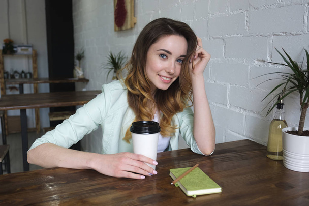 A beautiful young girl sits at a table in a big bright room with coffee in her hand and smiling looking into the camera. Horizontal photo - Foto, Imagen