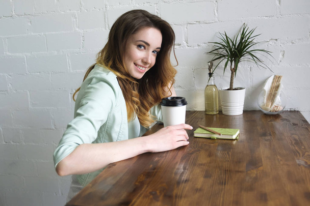 Beautiful young girl sitting at table with coffee and smiling looking at camera. Horizontal side photo - Zdjęcie, obraz