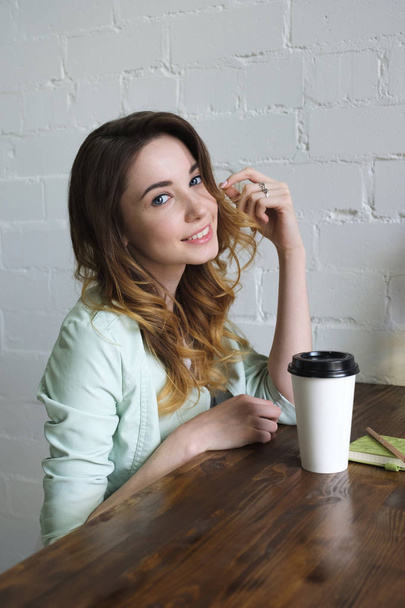 Hermosa joven se sienta en una mesa sobre un fondo de pared de ladrillo blanco con café en la mano y sonriendo mirando a la cámara. Foto lateral vertical
 - Foto, Imagen