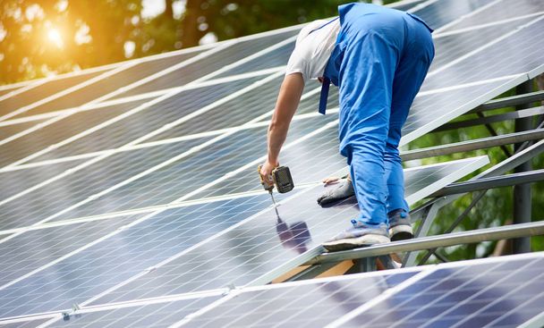 Construction worker connecting photo voltaic panel to solar system using screwdriver on shiny surface and lit by sun green tree background. Alternative energy and financial investment concept. - Photo, Image