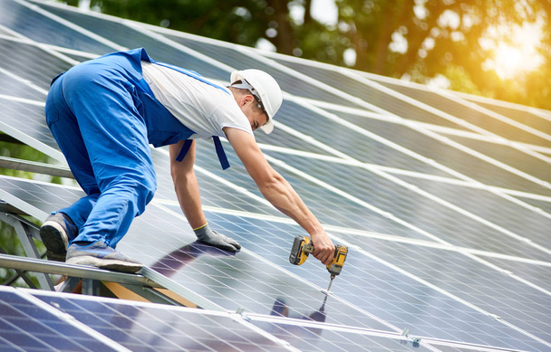 建設労働者は、光沢のある表面上のドライバーを使用して、太陽の緑の木の背景で点灯のソーラー システムに太陽光発電パネルを接続します。代替エネルギーと金融投資コンセプト. - 写真・画像