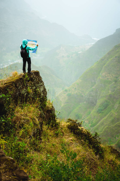 Hombre con el mapa que permanece en la cima de la roca frente a una magnífica vista panorámica de las altas cadenas montañosas y el exuberante valle verde
. - Foto, imagen