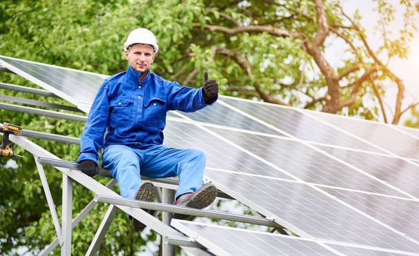 Der junge lächelnde Elektriker sitzt auf einer fast fertigen Photovoltaik-Solaranlage und zeigt die Daumen nach oben. auf sonnigem grünen Baumhintergrund. Alternatives Energiekonzept. - Foto, Bild