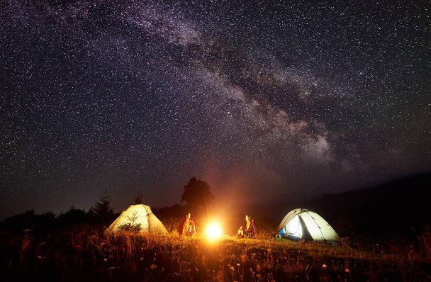 Night camping in mountains. Bright campfire burning between two backpackers, man and woman sitting opposite each other in front of illuminated tents under amazing dark blue starry sky and Milky way - Photo, Image