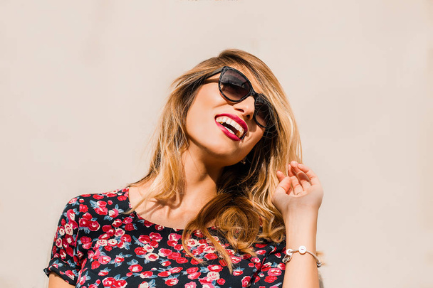Привлекательная молодая блондинка в солнечных очках и ярком платье, позирующем у стены
 - Фото, изображение