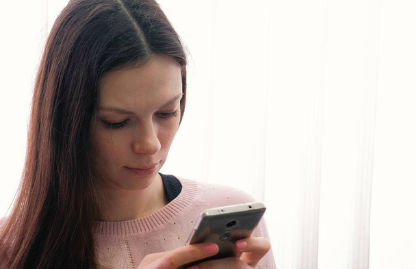 Young woman brunette is typing a message on her mobile phone and looking at screen. - Photo, Image