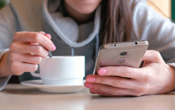 Woman is drinking a hot chocolate with spoon and look on the screen of mobile phone sitting in cafe. Closeup womans hands - Photo, Image