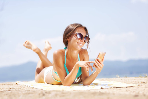 menina sexy com óculos de sol e um smartphone tomando banho de sol em uma praia
 - Foto, Imagem