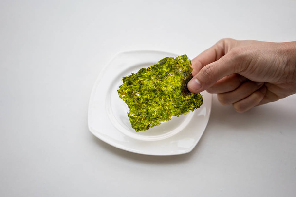 Mano recogiendo hoja de algas fritas coreanas sobre plato blanco sobre fondo blanco. Última pieza de muestra para pruebas
. - Foto, imagen