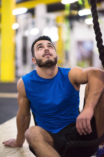 young muscular man sitting and relaxing before rope climbing in cross fitness gym - Valokuva, kuva