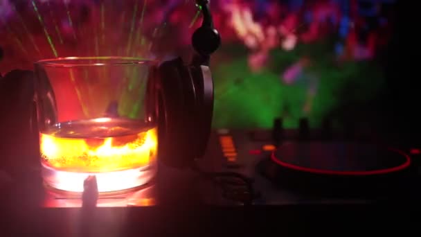 Glass with whisky with ice cube inside on dj controller at nightclub. Dj Console with club drink at music party in nightclub with disco lights. Selective focus - Felvétel, videó