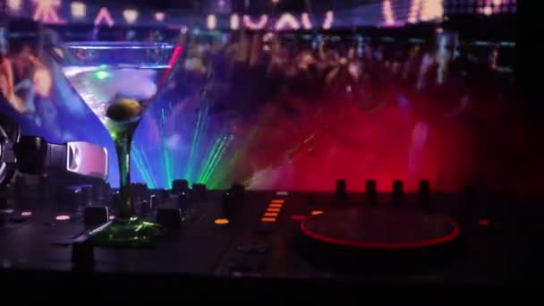 Glass with martini with olive inside on dj controller in night club. Dj Console with club drink at music party in nightclub with disco lights. - Footage, Video