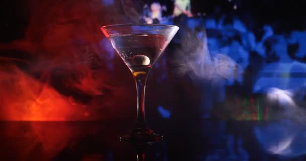 glas met martini met olijf erin. Close-up zicht op glas met club drankje op donkere mistige achtergrond. Selectieve focus. Clubdrankconcept - Video
