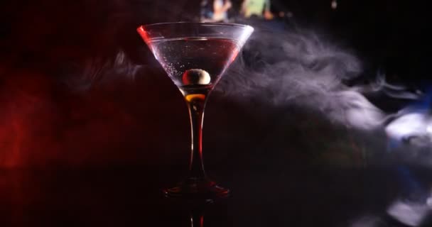 Glas mit Martini mit Oliven innen. Nahaufnahme des Glases mit Club-Drink auf dunklem, nebligem Hintergrund. Selektiver Fokus. Club-Drink-Konzept - Filmmaterial, Video