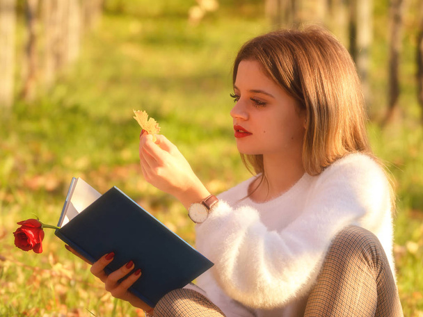 Girl reading a book sitting in the nature with a fallen leaf in her hand - Photo, Image