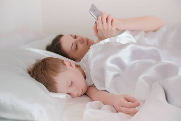 Mom woke up and browsing internet in her mobile phone. Her son is still asleep. - Foto, Imagem