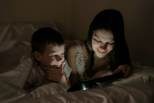 Mutter und Sohn entscheiden, was sie vor dem Schlafengehen auf dem Tablet unter der Decke sehen wollen. - Foto, Bild