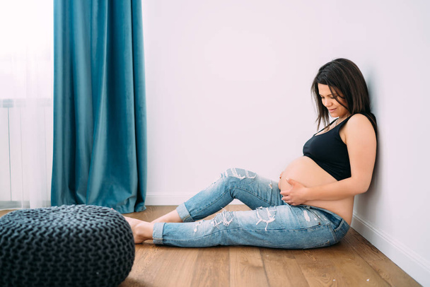happy smiling pregnant woman sitting on hardwood floor and touching her belly at new home - Fotografie, Obrázek
