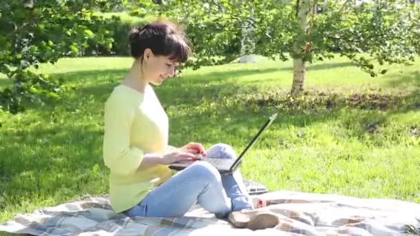Girl freelancer works in the open air.The brunette sits on the grass in the park and works with a laptop. Prints on the keyboard a message on the social network on the Internet. - Séquence, vidéo