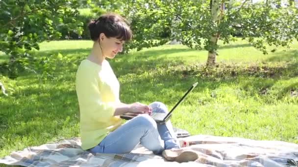 Girl freelancer works in the open air.The brunette sits on the grass in the park and works with a laptop. Prints on the keyboard a message on the social network on the Internet. - Video, Çekim