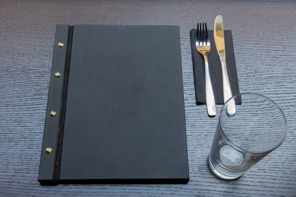 Чорне або темно-сіре меню ресторану з копіювальним простором, виделкою та ножем та порожнім склом на темному дерев'яному столі
 - Фото, зображення