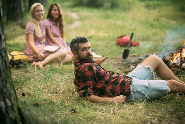 Bearded man in lumberjack shirt lying on grass, unity with nature concept. Group of young hikers camping in forest - Foto, Imagem