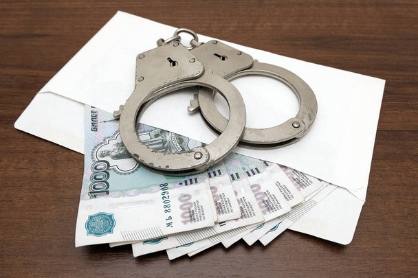 Een bundel van Russische roebels in een envelop en handboeien op een bruine achtergrond. Het concept van overtreding van de wet in het financiële gebied, fraude, corruptie, omkoping. - Foto, afbeelding