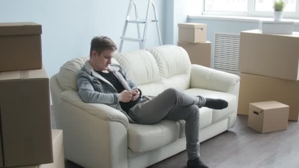 El joven se mudó a un nuevo apartamento. Se sienta en el sofá con un teléfono inteligente
 - Metraje, vídeo