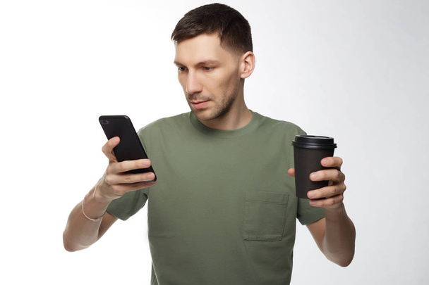 Thoughtful serious brunette male entrepreneur in khaki t-shirt, has coffee break after hard work, makes business report by modern digital phone, free internet connection, waits for call from partner. - Photo, Image
