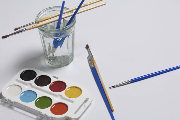 Watercolors and brushes on a white surface. Next glass for water. Subjects for creativity. - Foto, imagen