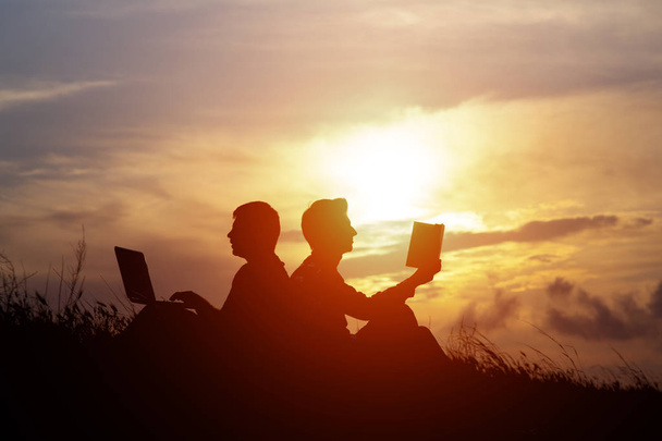 силует чоловіків з ноутбуком і книгою на заході сонця або на фоні сходу сонця. Ідея протиріч старих і нових цінностей. Поняття старого і сучасного
 - Фото, зображення