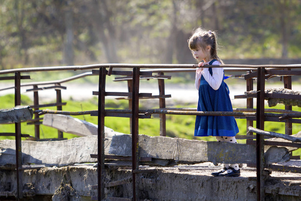 Melko pieni vaalea pitkäkarvainen tyttö mukava sininen mekko seisoo yksin vanha sementti silta nojaten puiset kaiteet etsivät intensiivisesti alas lämmin aurinkoinen päivä. Lapset uteliaisuus ja fantasiat käsite
. - Valokuva, kuva