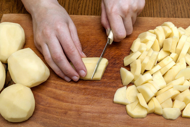 Женская рука с ножом, резающим картофель на деревянной доске на кухне. Здоровое питание и образ жизни. Вид сверху крупным планом
 - Фото, изображение