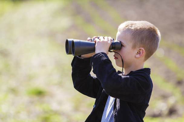 Portrait in profile of little handsome cute blond boy looking thoughtfully through binoculars in distance on blurred background. Children dreams, fantasies and imaginations concept. - Photo, Image