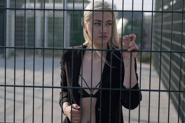menina loira em um casaco preto atrás de uma cerca de metal
 - Foto, Imagem