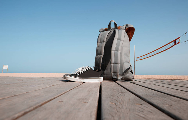 Summer vacation concept. Backpack and sneakers are on the wooden desk near the beach and sea.Blue clear sky and sun. - Photo, image