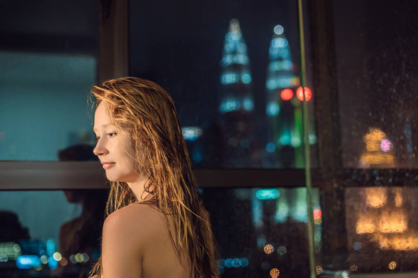 Beautiful woman wipes her hair after shower on background of window with panoramic view of night city. - Foto, Imagen